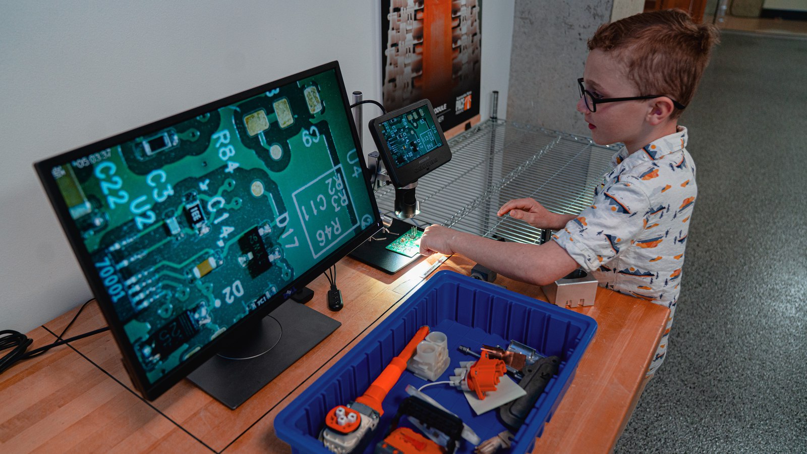 Boy in glasses steering a circuit board under a digital microscope, magnified on a big monitor