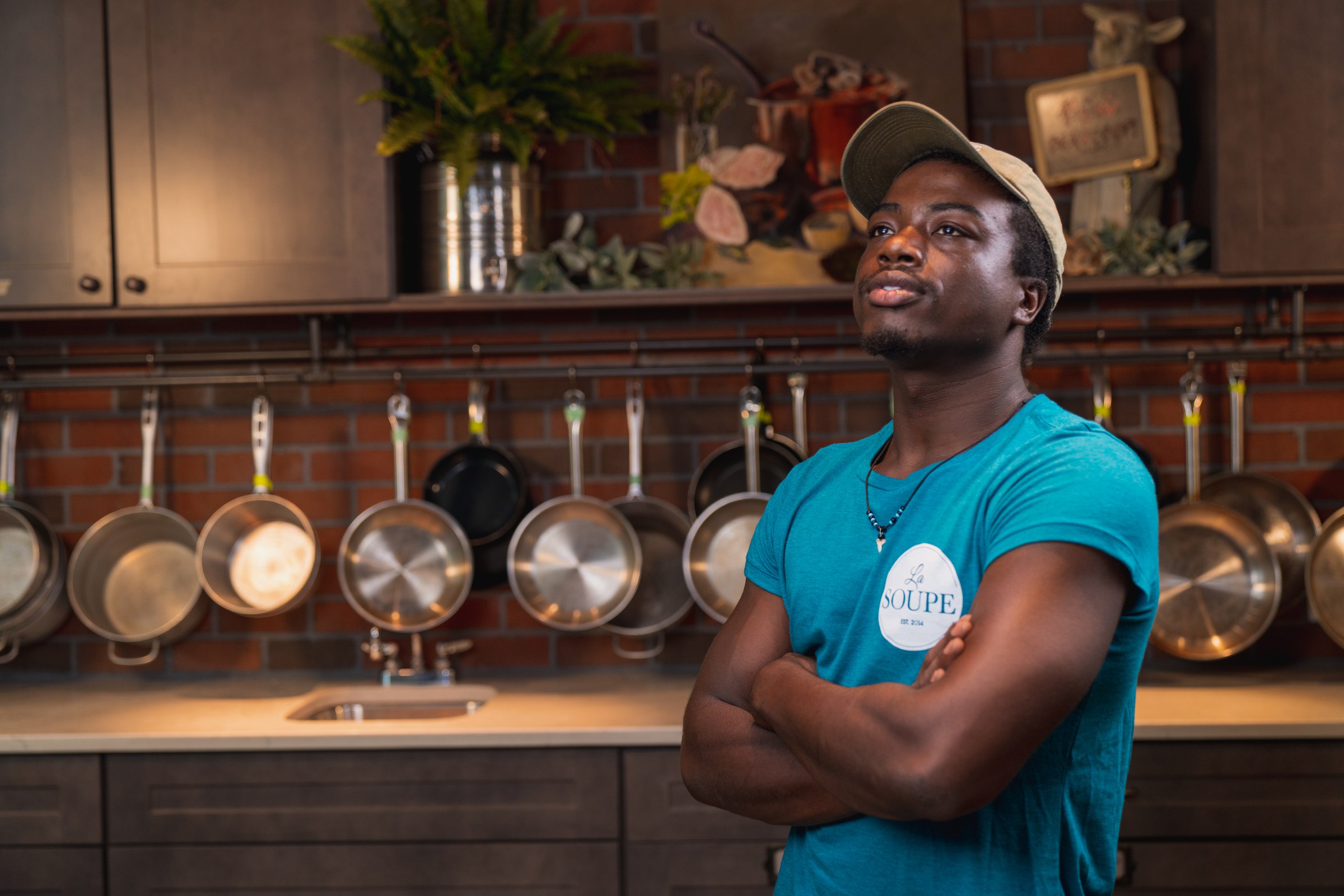 Cook in a teal La Soupe shirt, arms crossed, in front of a wall of hanging pans