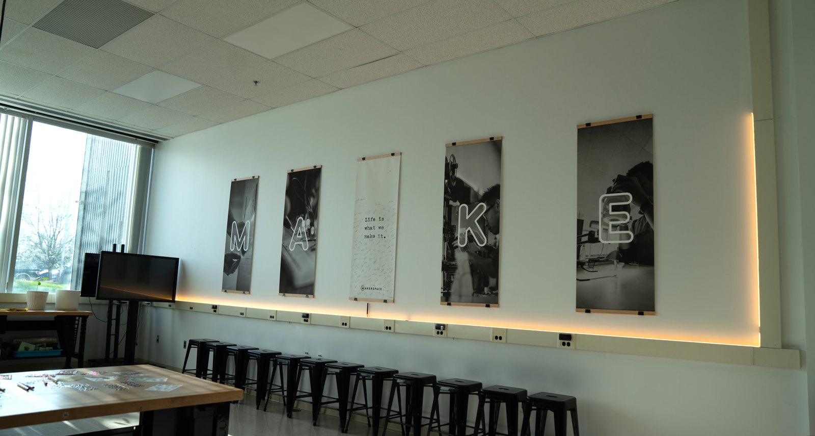 Five tall black-and-white banners spelling MAKE above a row of stools in the Workshop