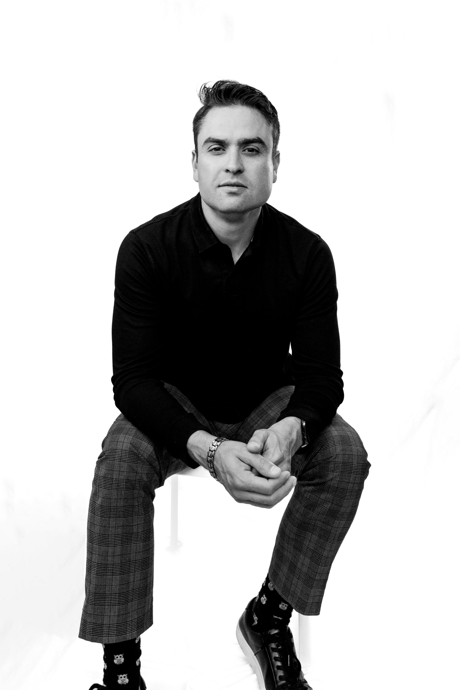 Black and white portrait of a man seated on a stool in a black polo, plaid trousers and owl socks