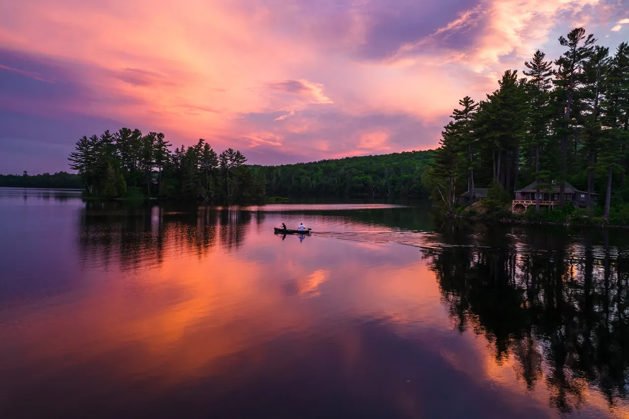 A lake at sunset, purple and orange sky mirrored in still water beside pines