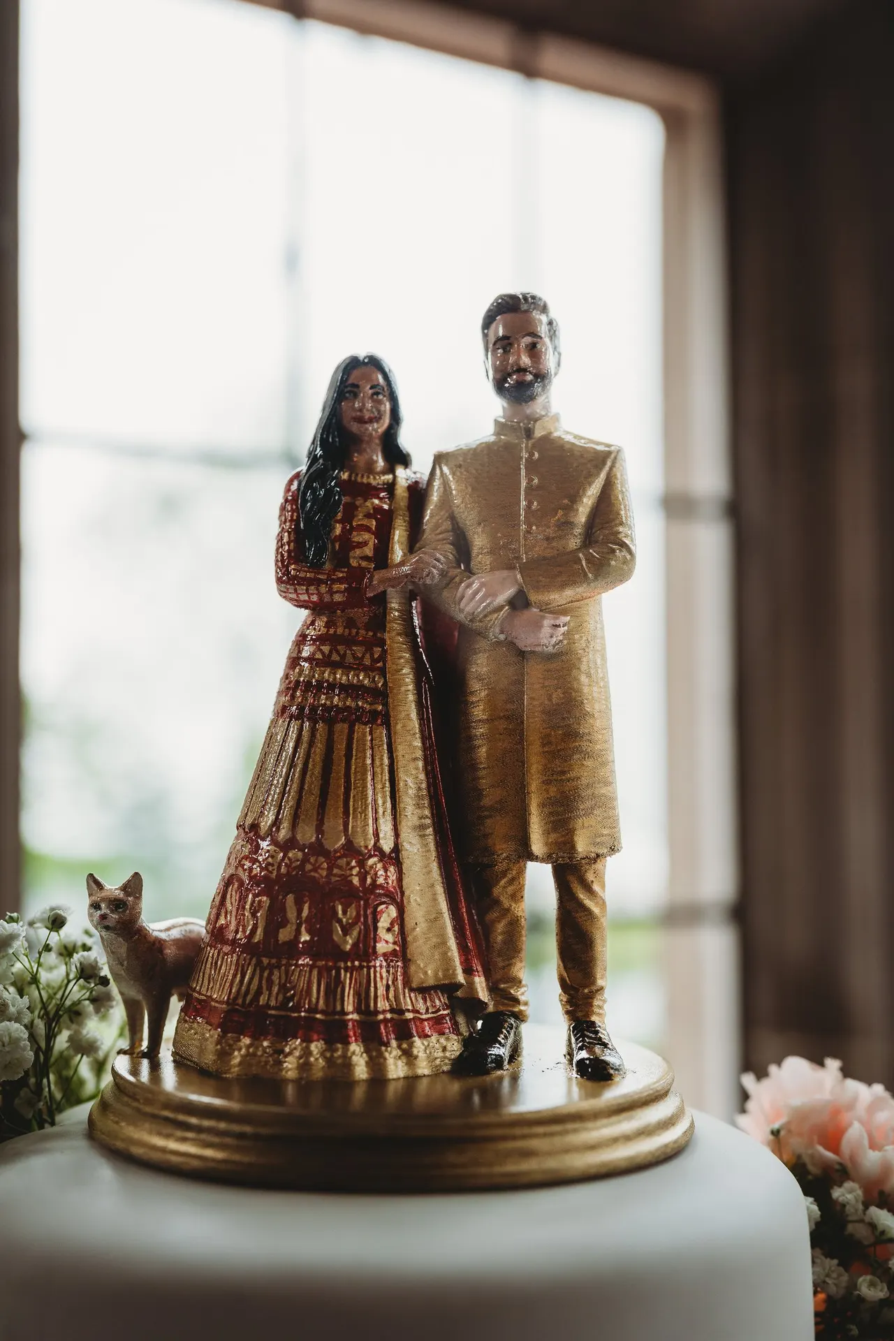 A painted figurine of a couple in wedding clothes with their cat, on a gold base on top of a white cake
