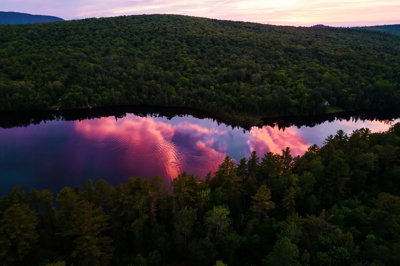 Pink sunset clouds reflected in a lake between forested hills, from the drone