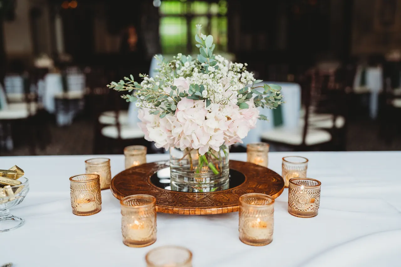 An engraved wooden ring with a mirror, a vase of pink and white flowers and gold votive candles on a round table