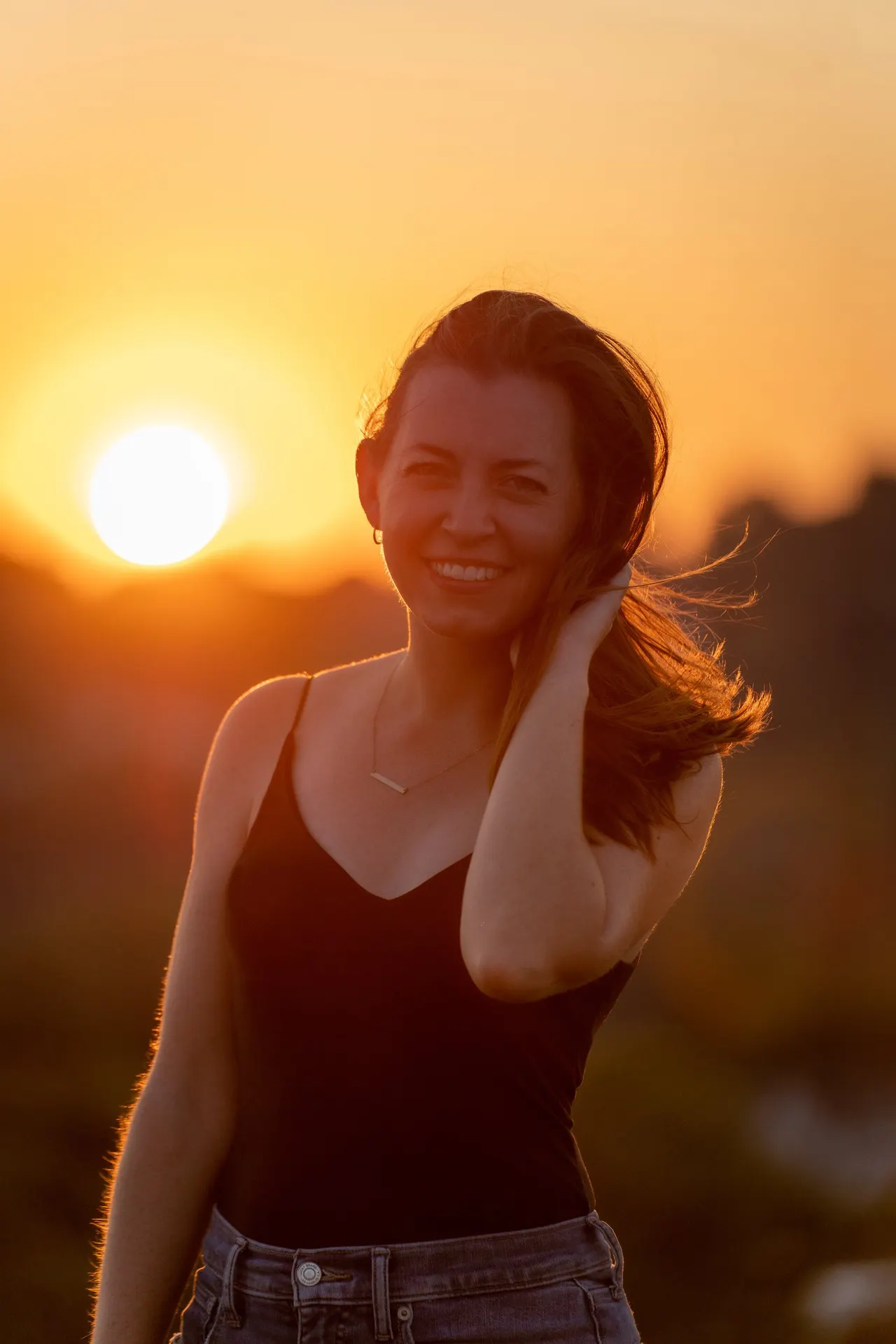A woman smiling at sunset, the sun glowing beside her over the city's hills