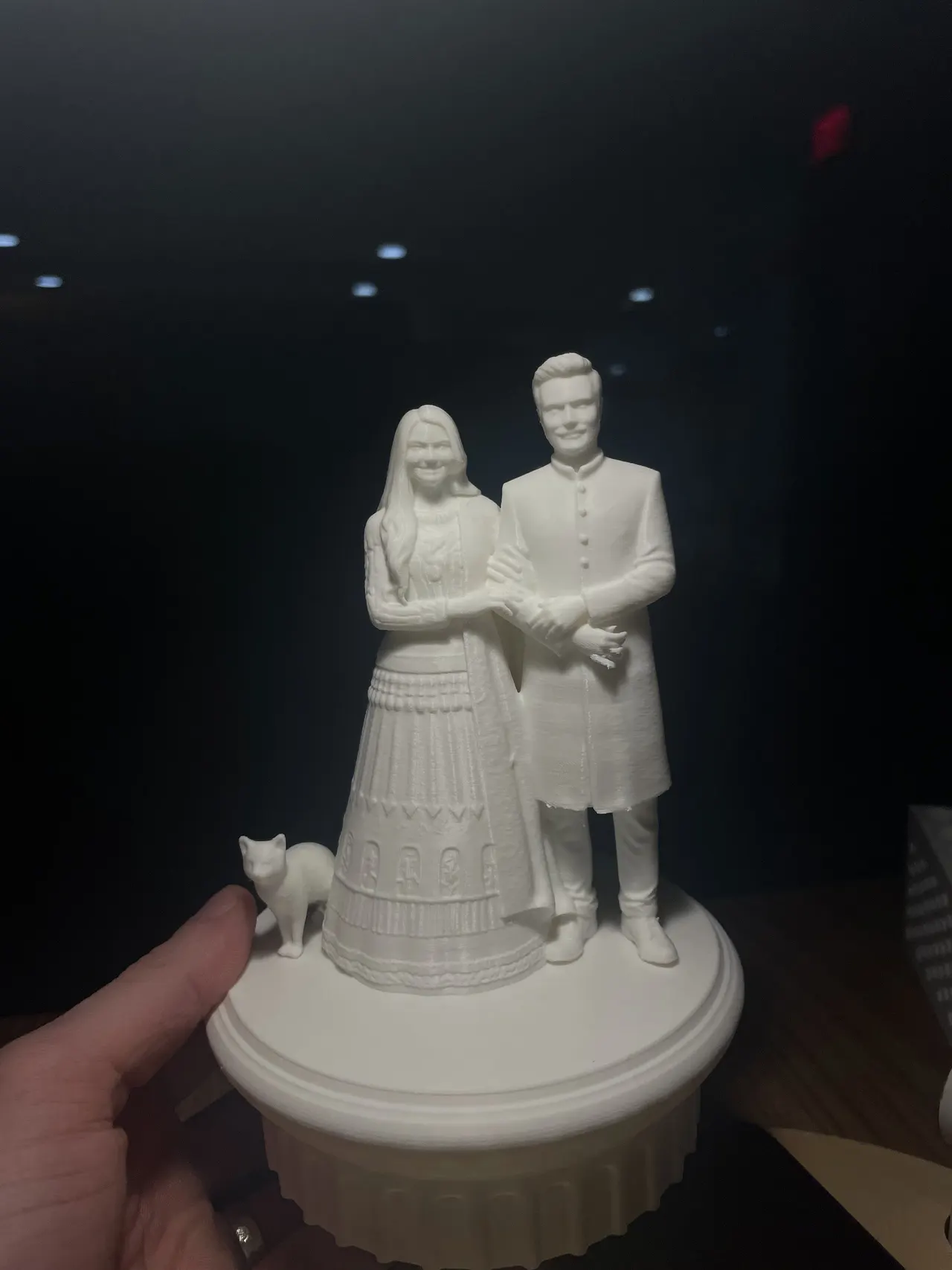 A white 3D print of a couple in wedding clothes with their cat, held up in a hand