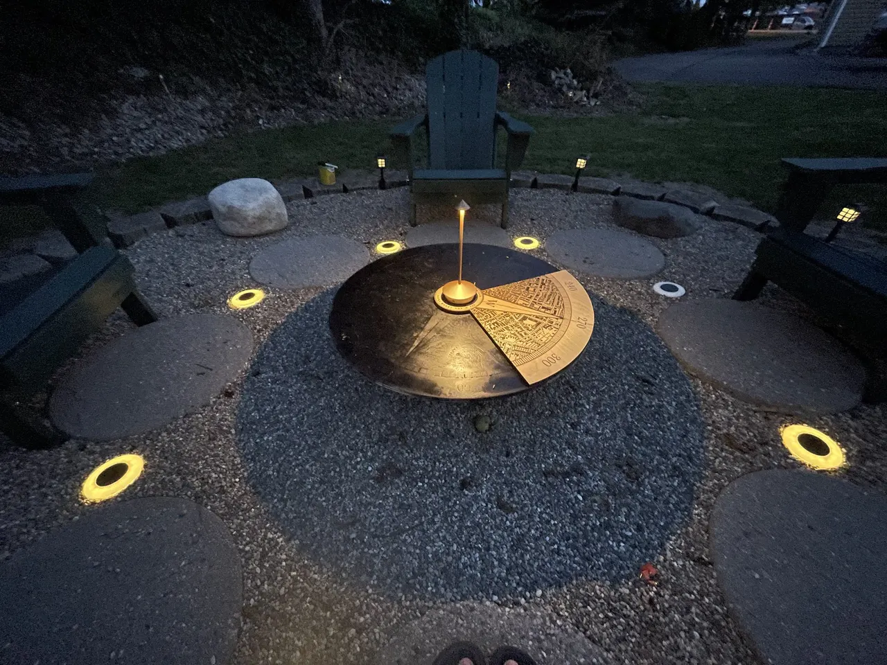 A backyard fire pit at dusk with the compass topper in the middle, ringed by lights and chairs