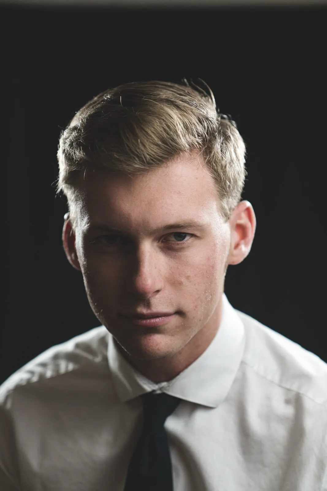 A young man in a white shirt and tie, lit against black